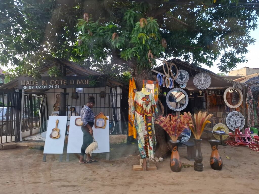 Scène animée de la rue des artisans à Grand-Bassam, bordée d’échoppes colorées proposant des souvenirs traditionnels : sculptures en bois, bijoux, tissus imprimés et objets décoratifs.