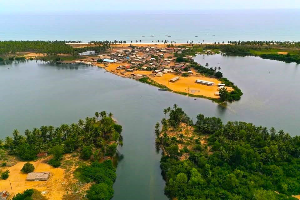 Image panoramique capturant la ville côtière de Grand-Lahou en Côte d’Ivoire. Au premier plan, la lagune Tagba serpente entre les terres, tandis qu’au fond, l’océan Atlantique s’étend sous un ciel dégagé. Ce paysage met en valeur la rencontre entre lagune, mer et territoire urbain.