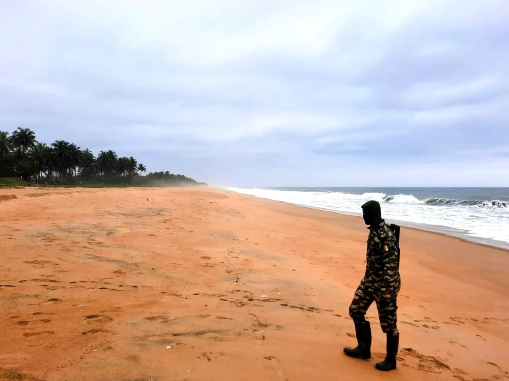 Image représentant un guide de l’Office Ivoirien des Parcs et Réserves (OIPR) marchant sur le sable fin de Lahou Plage. En arrière-plan, l’océan Atlantique s’étend à perte de vue sous un ciel avec des nuages.