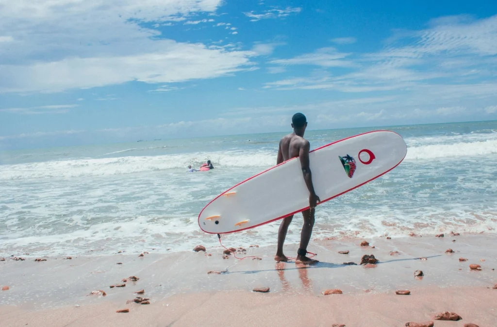 Surfeur sur la plage en Côte d’Ivoire avec une planche de surf sous le bras, devant la mer et un ciel bleu clair.