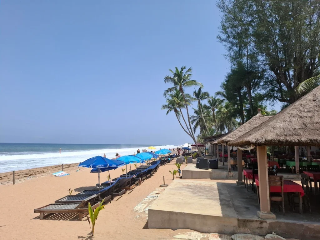 Plage d’Assinie en Côte d’Ivoire avec parasols alignés, sable doré, cocotiers et mer calme sous un ciel bleu.
