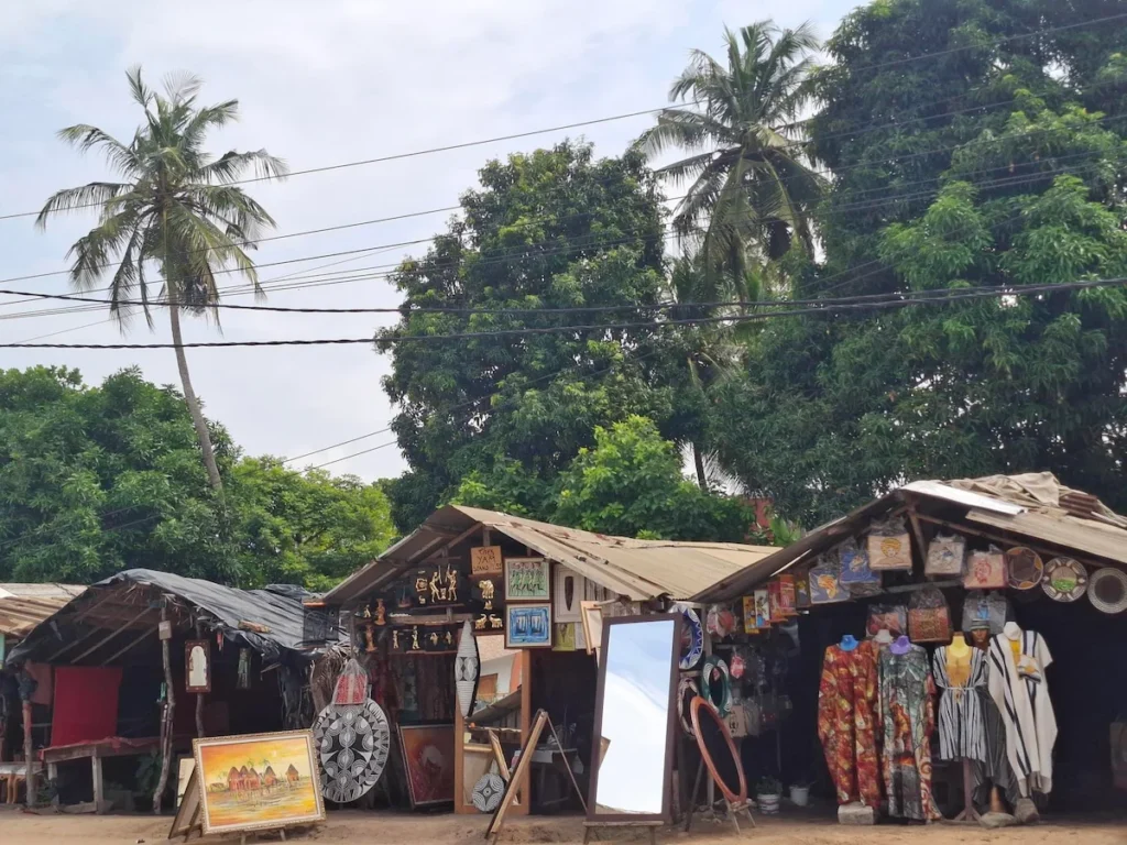 Visiter-Grand-Bassam-Cote-dIvoire-plages-culture-hotel-rue-artisans Boutiques d’artisans à Grand-Bassam, avec étals de sculptures et objets artisanaux devant la rue.