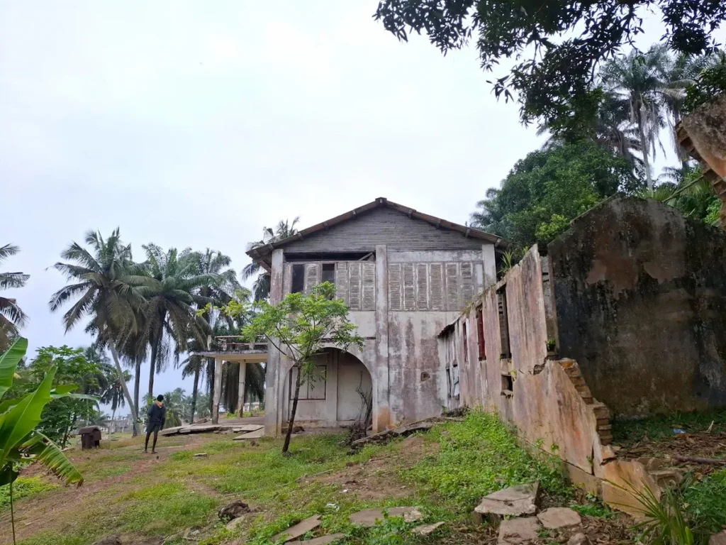 Façade de la Maison des Esclaves de Sassandra, un bâtiment ancien en pierre entouré de palmiers et de végétation.