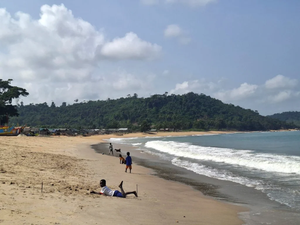 Enfants jouant au bord de la plage de Monogaga, avec sable doré, vagues puissantes et collines verdoyantes en arrière-plan.