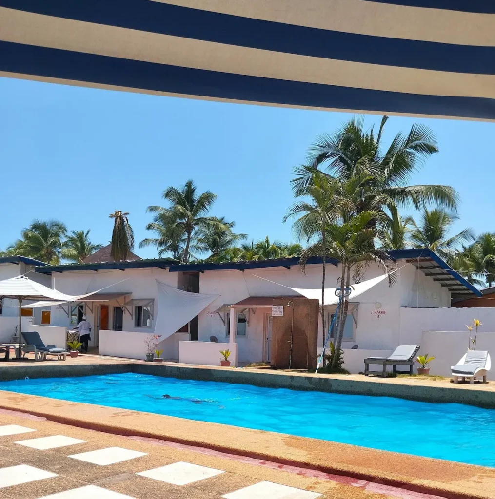 Visiter-Grand-Bassam-Cote-dIvoire-plages-culture-hotel-madrague Piscine extérieure entourée de chambres et palmiers à Grand-Bassam, sous un ciel clair.