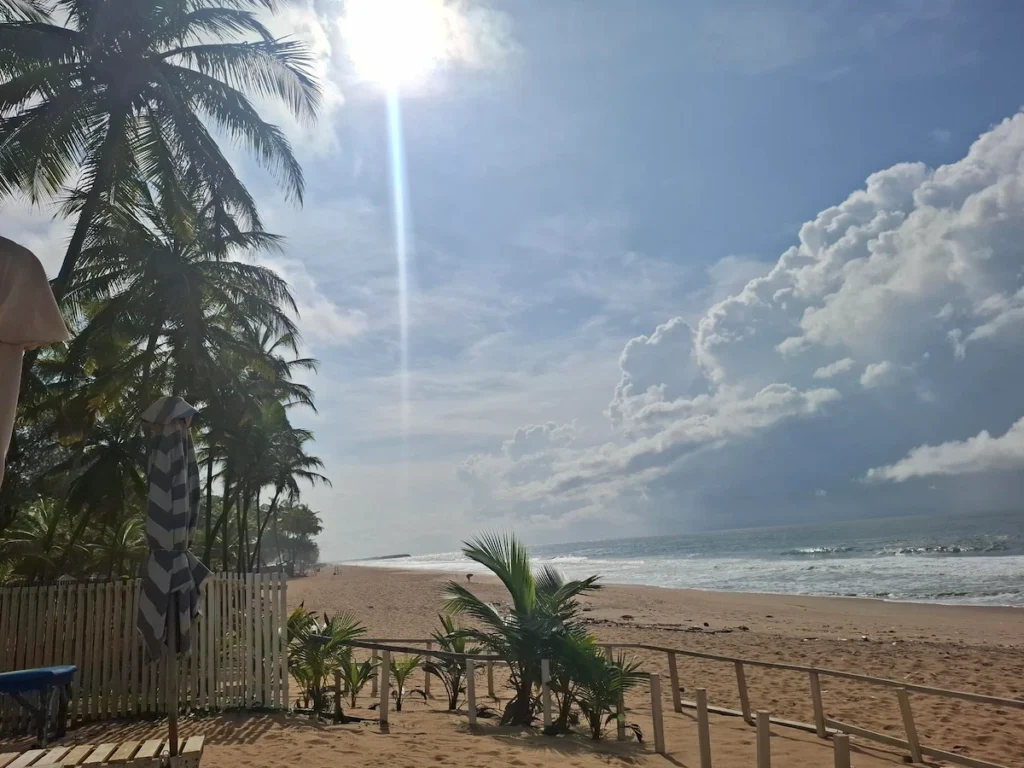 Visiter-Grand-Bassam-Cote-dIvoire-plages-culture-hotel Plage ensoleillée à Grand-Bassam en Côte d’Ivoire, bordée de cocotiers sous un ciel bleu lumineux.