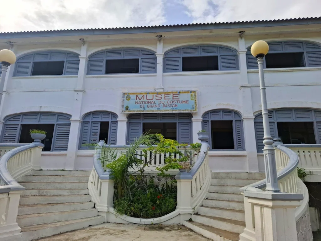 Visiter-Grand-Bassam-Cote-dIvoire-plages-culture-hotel-quartier-france-musee-national-costume Façade du Musée National du Costume à Grand-Bassam, avec escaliers blancs et végétation à l’entrée.