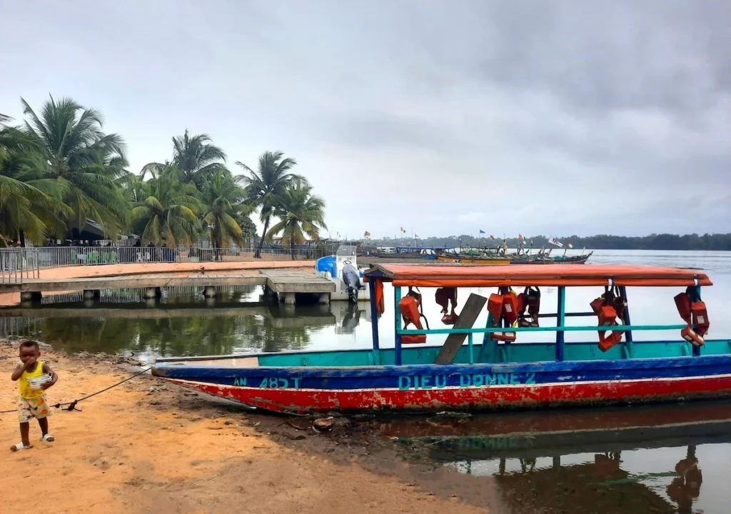 Bateaux colorés amarrés sur la lagune à Grand-Lahou, avec un enfant debout sur le rivage près des palmiers. 