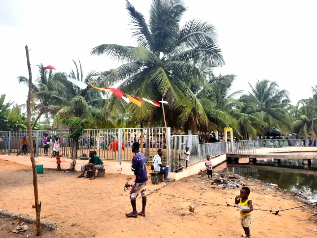 Enfants jouant sur une place sableuse à Grand-Lahou, avec palmiers et bâtiments en arrière‑plan. 