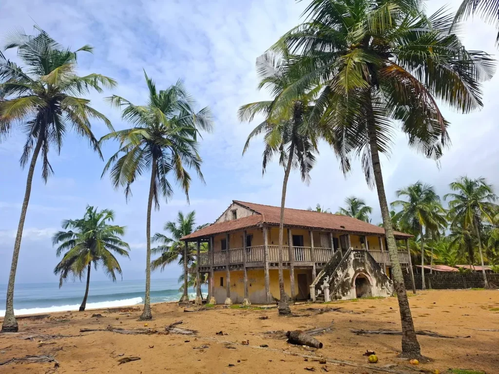 Maison coloniale au bord de la plage de Jacqueville, entourée de palmiers et sable doré sous un ciel clair.