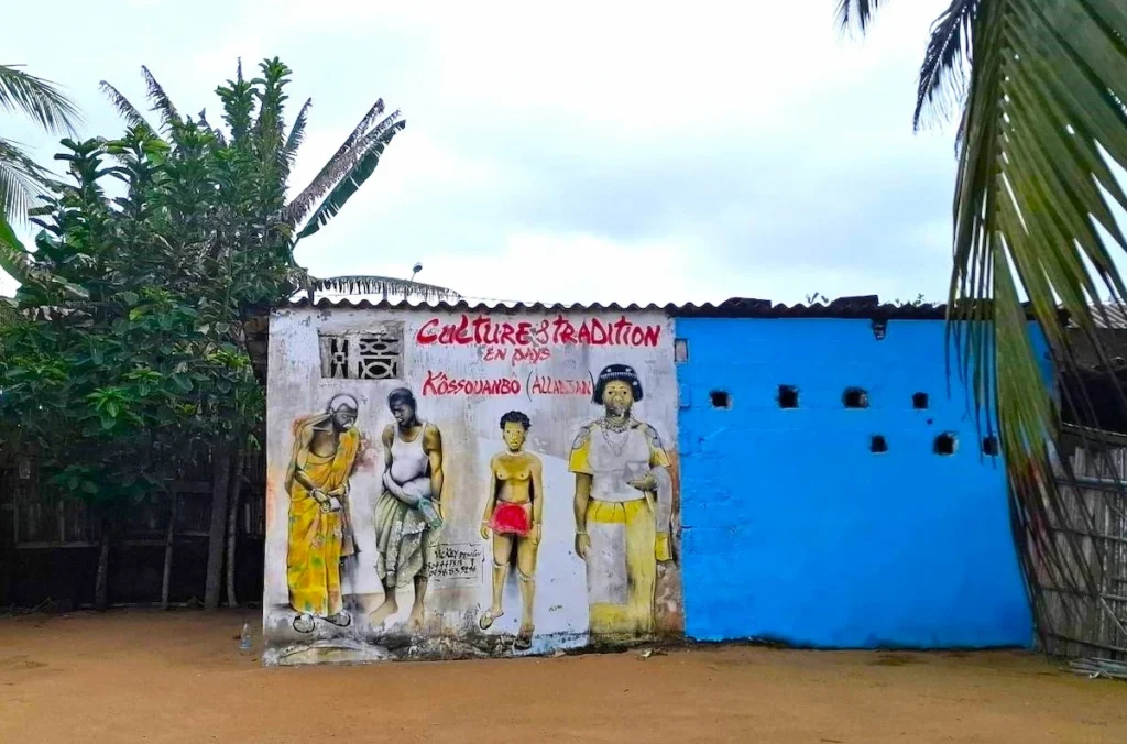 Peinture murale représentant les ethnies Alladian à Jacqueville, entourée de palmiers et d’un bâtiment coloré.