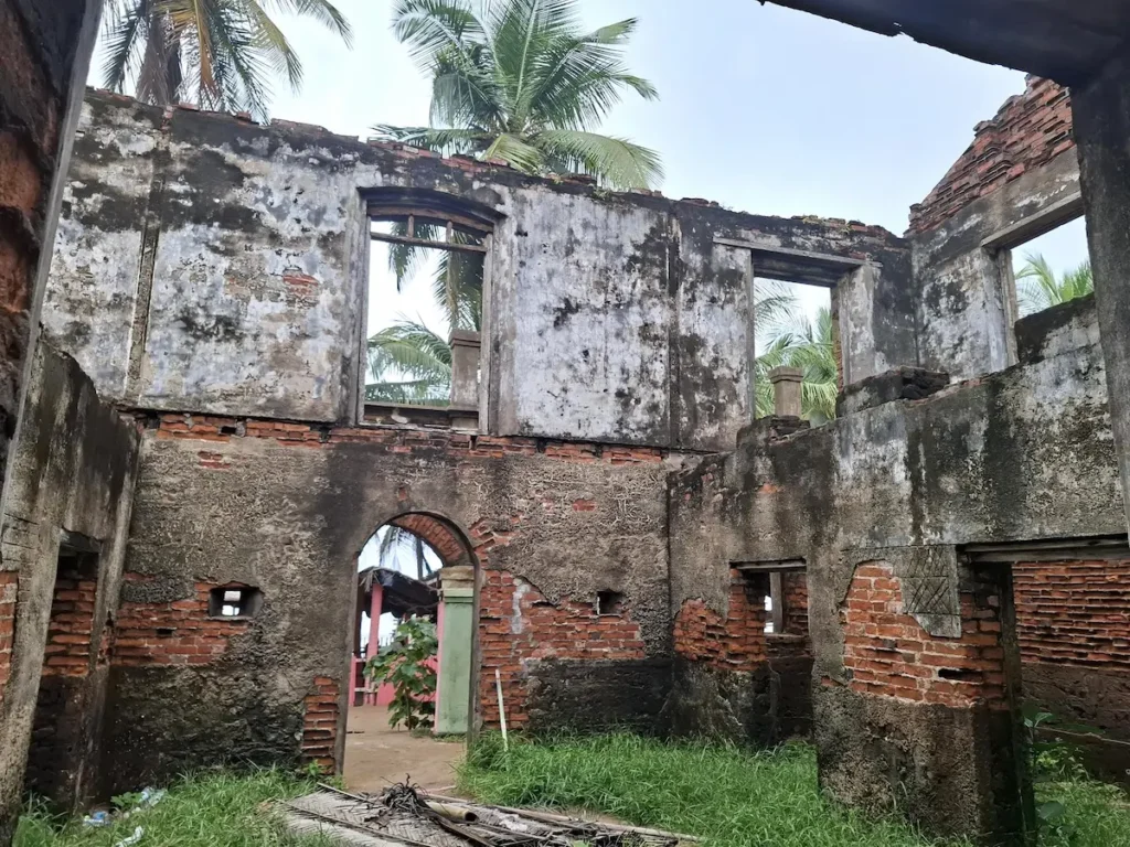 Intérieur en ruines du maquis Papa John à Jacqueville, avec murs ouverts sur l’extérieur et palmiers visibles au loin.