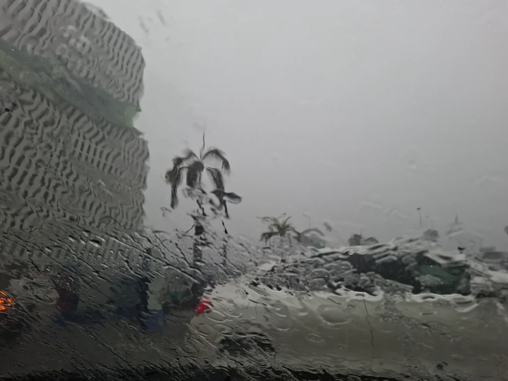 Forte pluie à Abidjan vue depuis l’intérieur d’une voiture, avec route mouillée et palmiers sous un ciel gris. 